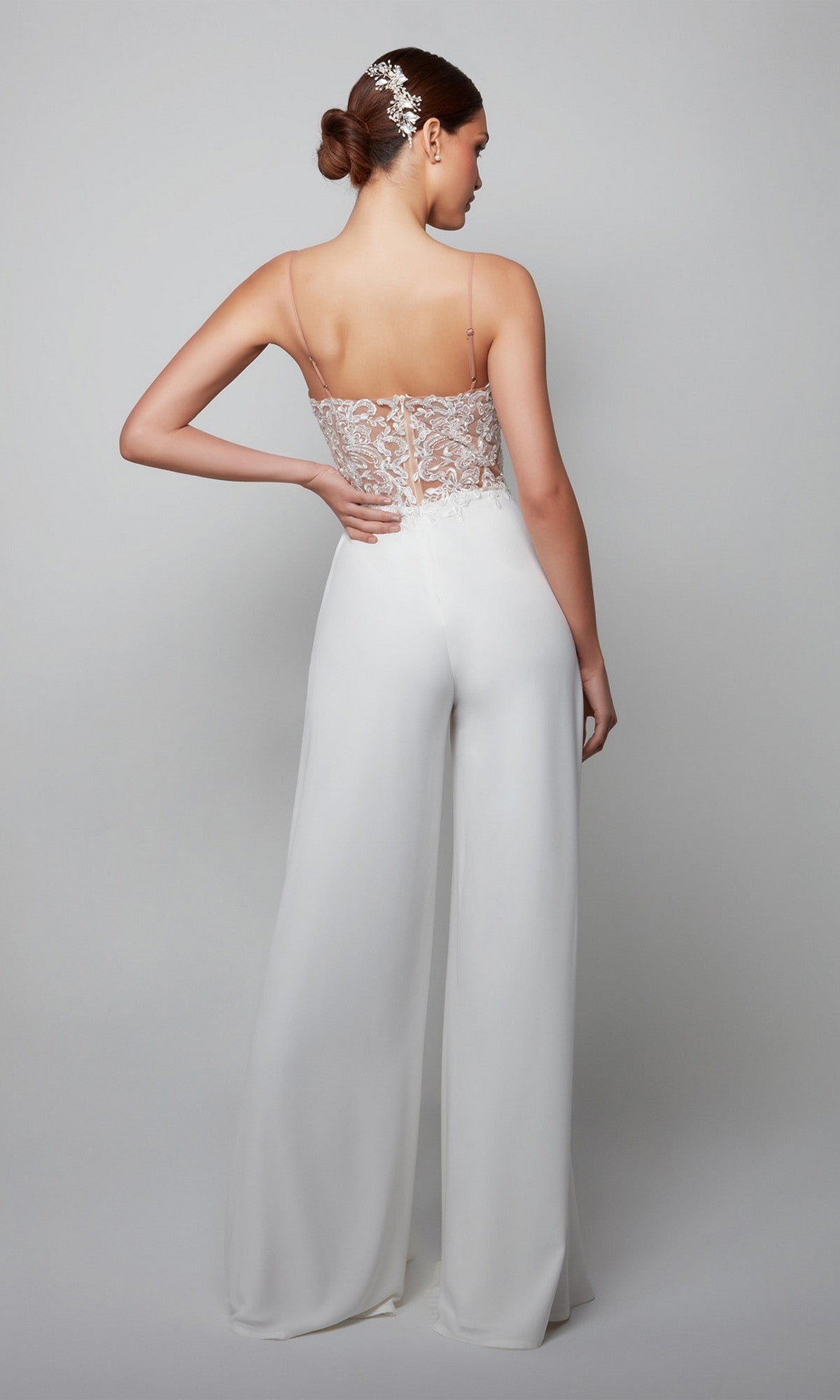 Woman facing away in the Long White Jumpsuit: Alyce 70017, which has a white lace bodice, backless design with spaghetti straps, and wide-leg pants. Her hair is styled in a bun with a decorative accessory.
